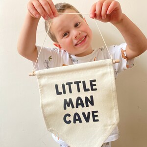 May include: A white canvas banner with black text that reads "LITTLE MAN CAVE". A young person is holding the banner up in front of them.