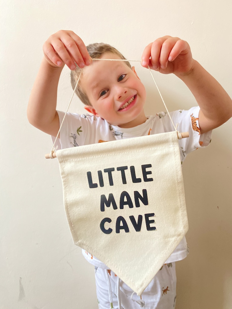 May include: A white canvas banner with black text that reads "LITTLE MAN CAVE". A young person is holding the banner up in front of them.
