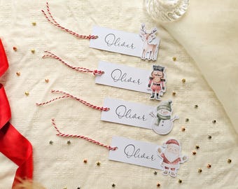 May include: Four rectangular white gift tags with red and white baker's twine. Each tag features the name Oliver in black script and a Christmas-themed illustration: a reindeer, a nutcracker, a snowman, and a Santa Claus figure.