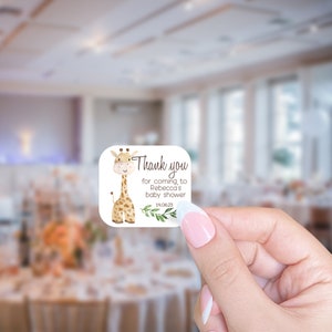 May include: Thank you for coming to Rebecca's baby shower 14.06.23. A white square sticker with a cartoon giraffe illustration and green leaves.