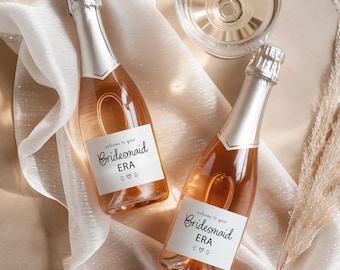 May include: Two glass bottles of rose wine with silver foil caps, each featuring a white rectangular label with the black text "welcome to your Bridesmaid ERA" and a small heart icon. The bottles are positioned on a sheer white fabric.