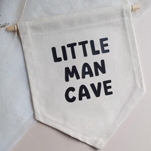 May include: A white canvas banner with black text that reads "Little Man Cave". The banner is hanging from a string and has a wooden dowel at the top.