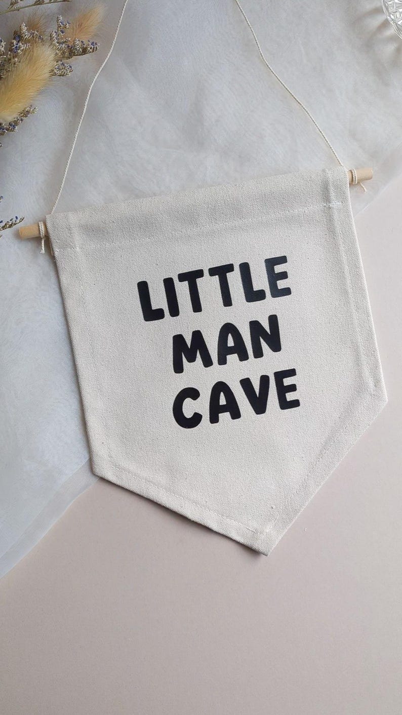 May include: A white canvas banner with black text that reads "Little Man Cave". The banner is hanging from a string and has a wooden dowel at the top.