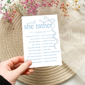 以下が含まれることがあります： 青と白の印刷可能なゲームカードで、「would she rather」というテキストと、花嫁に尋ねる質問のリストが書かれています。カードは薄い茶色の表面の上にあります。