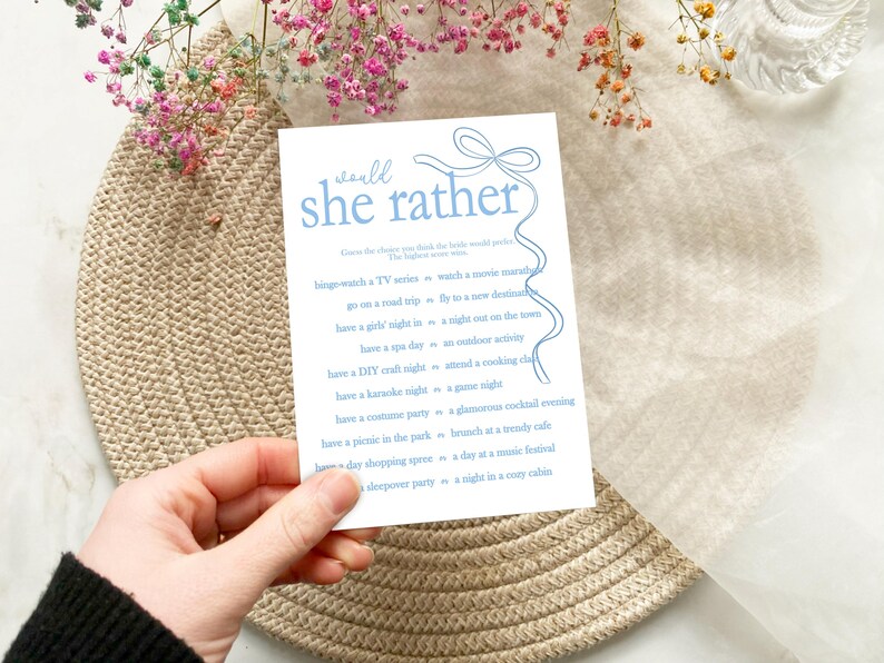 以下が含まれることがあります： 青と白の印刷可能なゲームカードで、「would she rather」というテキストと、花嫁に尋ねる質問のリストが書かれています。カードは薄い茶色の表面の上にあります。