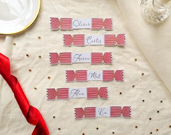 May include: Six paper place cards shaped like Christmas crackers, each featuring a pink and white zigzag pattern on the ends and a white center panel printed with the names Oliver, Curtis, Aaron, Mat, Alex, and Kim in black script.