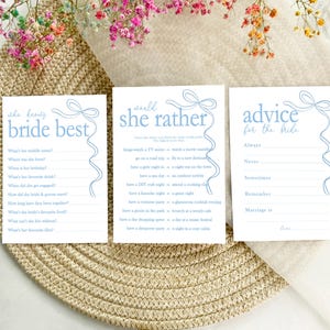 以下が含まれることがあります： 青いリボンをあしらった、印刷可能なブライダルシャワーゲームカード3枚。最初のカードは「She Knows Bride Best」というタイトルで、花嫁に関する質問が書かれています。2枚目のカードは「She'd Rather」というタイトルで、花嫁のための選択肢がリストされています。3枚目のカードは「Advice for the Bride」というタイトルで、ゲストがアドバイスを与えるためのプロンプトがリストされています。