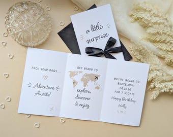 May include: A white tri-fold greeting card with black text and graphics. The front reads a little surprise with a black ribbon. Inside, it says PACK YOUR BAGS, Adventure Awaits, GET READY TO explore, discover & enjoy with a world map, and YOU'RE GOING TO BARCELONA! 06.10.26 FOR 7 NIGHTS, Happy Birthday Holly.
