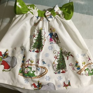 Puede incluir: Un vestido blanco con un lazo verde en el cuello. El vestido tiene un estampado navideño con un tren, árboles y personajes. El vestido tiene dos bolsillos a los lados.