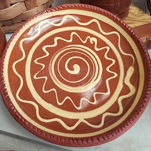 May include: A handmade, terracotta-colored ceramic plate with a cream-colored spiral and wavy design. The plate has a raised rim and is likely used for serving food. The design is centered on the plate and is a decorative element.