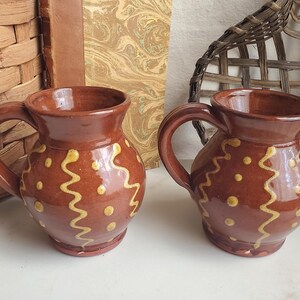 May include: Two matching brown ceramic mugs with a glossy finish, each featuring a rounded body, a curved handle, and a flared rim. The exterior of each mug is decorated with vertical yellow wavy lines and small yellow dots painted in a repeating pattern.