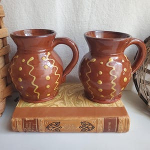 May include: Two matching brown ceramic mugs with a glossy finish, each featuring a rounded body, a curved handle, and a flared rim. The exterior of each mug is decorated with vertical yellow wavy lines and small yellow dots in a repeating pattern.