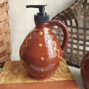 May include: A brown ceramic soap dispenser with a black pump and handle. The dispenser is decorated with gold wavy lines and dots. It sits on a decorative surface, with woven baskets in the background.