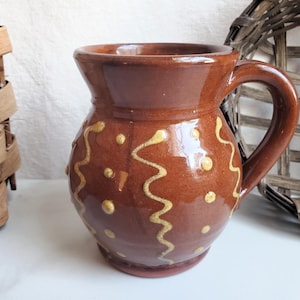May include: A glossy brown ceramic mug with a rounded body and a single handle. The exterior features a hand-painted decorative pattern consisting of vertical wavy lines and small dots in a light cream color, applied over the smooth brown glaze.