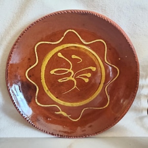 May include: A handmade, terracotta-colored ceramic plate with a yellow, stylized sun design in the center. The plate has a scalloped edge and is likely used for serving food. The plate is a unique piece of pottery.