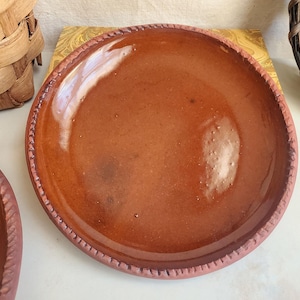 May include: A handmade, terracotta-colored plate with a textured rim. The plate has a smooth, glossy surface and a slightly raised edge. The plate is a warm, earthy tone, perfect for serving food.