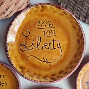 May include: A round, ceramic plate with a yellow-brown glaze and a dark brown rim. The plate features the word "Liberty" in cursive, with the numbers "1776" and "1781" above it. The plate has a crackled texture.