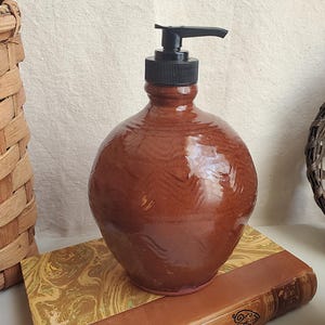 May include: A brown ceramic soap dispenser with a black pump top. The dispenser has a rounded shape and sits on a book with decorative marbled paper. Wicker baskets are in the background.