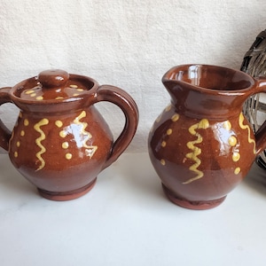 May include: A brown ceramic sugar bowl with a lid and a matching creamer. Both pieces are decorated with yellow wavy lines and dots. The sugar bowl has two handles. The creamer has a handle and a spout. The set is likely vintage.
