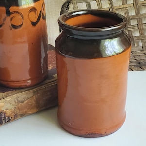 May include: Two ceramic jars with a rustic, vintage aesthetic. The jars are a warm brown color with a dark brown rim and accents. One jar has a handle and the other has decorative swirls. The jars are sitting on a book.