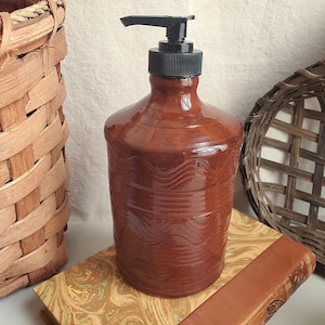 May include: A brown ceramic soap dispenser with a black pump. The dispenser has a glossy finish and a wavy design around the middle. It is sitting on a decorative book with a woven basket in the background.