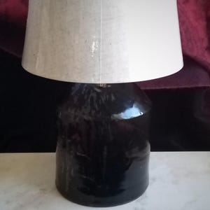 May include: A black ceramic table lamp with a white lampshade. The lamp has a glossy finish and a rounded base. The lampshade is cylindrical and made of a light-colored fabric. The lamp is sitting on a white surface.