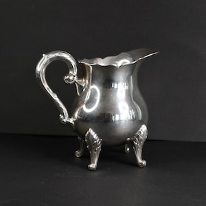 May include: A silver creamer with a decorative handle and three ornate feet. The creamer has a scalloped rim and a curved spout. The handle features a scroll design. The creamer is made of a shiny metal.