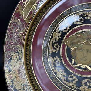 Op de afbeelding: Close-up van een decoratieve plaat met een bordeauxrode, zwarte en gouden kleurstelling. De plaat heeft sierlijke gouden details, waaronder een centraal Medusa-hoofdontwerp en ingewikkelde krulpatronen. Het ontwerp van de plaat is symmetrisch en elegant.