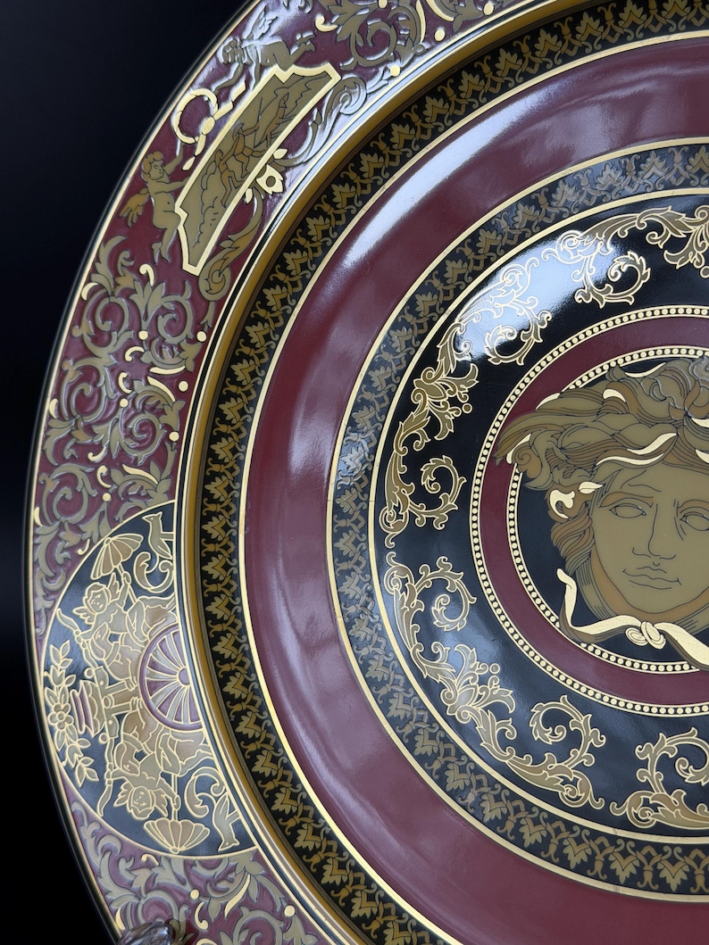 Op de afbeelding: Close-up van een decoratieve plaat met een bordeauxrode, zwarte en gouden kleurstelling. De plaat heeft sierlijke gouden details, waaronder een centraal Medusa-hoofdontwerp en ingewikkelde krulpatronen. Het ontwerp van de plaat is symmetrisch en elegant.