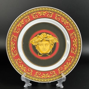 Puede incluir: Un plato decorativo con una cabeza de Medusa dorada en el centro, rodeado por un borde rojo y dorado ornamentado. El plato tiene un centro negro y un anillo interior blanco. El borde exterior presenta un dise&ntilde;o de volutas doradas.