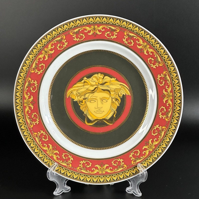 Puede incluir: Un plato decorativo con una cabeza de Medusa dorada en el centro, rodeado por un borde rojo y dorado ornamentado. El plato tiene un centro negro y un anillo interior blanco. El borde exterior presenta un dise&ntilde;o de volutas doradas.