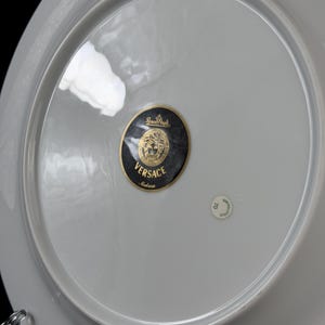 Op de afbeelding: Een witte porseleinen bord met een grijze rand. De plaat heeft een zwarte en gouden Versace Medusa logo sticker in het midden. Een kleine, ronde sticker is ook zichtbaar. De plaat maakt waarschijnlijk deel uit van een servies.