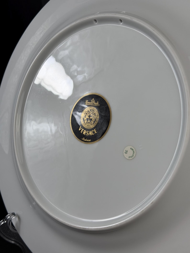 Op de afbeelding: Een witte porseleinen bord met een grijze rand. De plaat heeft een zwarte en gouden Versace Medusa logo sticker in het midden. Een kleine, ronde sticker is ook zichtbaar. De plaat maakt waarschijnlijk deel uit van een servies.