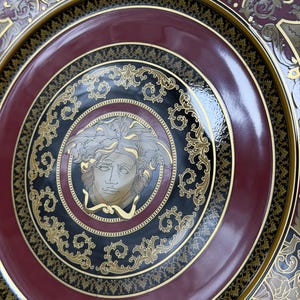 Op de afbeelding: Decoratieve plaat met een bordeauxrode, zwarte en gouden kleurstelling. De plaat heeft een centraal Medusa-hoofdontwerp, omgeven door sierlijke gouden krullen en een glanzende afwerking. De rand heeft een gedetailleerd patroon.