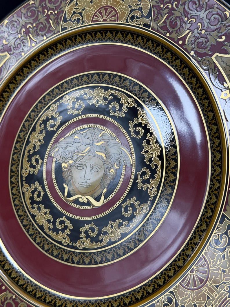 Op de afbeelding: Decoratieve plaat met een bordeauxrode, zwarte en gouden kleurstelling. De plaat heeft een centraal Medusa-hoofdontwerp, omgeven door sierlijke gouden krullen en een glanzende afwerking. De rand heeft een gedetailleerd patroon.