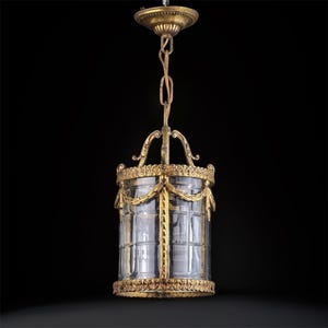 Puede incluir: Una lámpara colgante antigua de color dorado con una pantalla de vidrio transparente y rectangular. La lámpara tiene detalles ornamentados, incluyendo guirnaldas decorativas y una parte superior circular. La luz cuelga de una cadena y un soporte de techo decorativo.