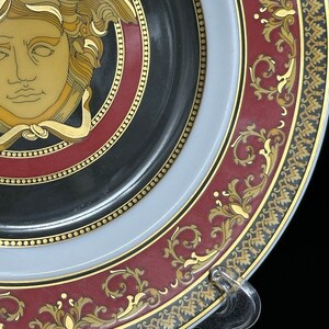 Puede incluir: Plato decorativo con un dise&ntilde;o de cabeza de Medusa en oro y marr&oacute;n sobre un fondo negro. El plato presenta un borde burdeos con adornos dorados y un borde gris claro. Un soporte de pl&aacute;stico transparente sostiene el plato.