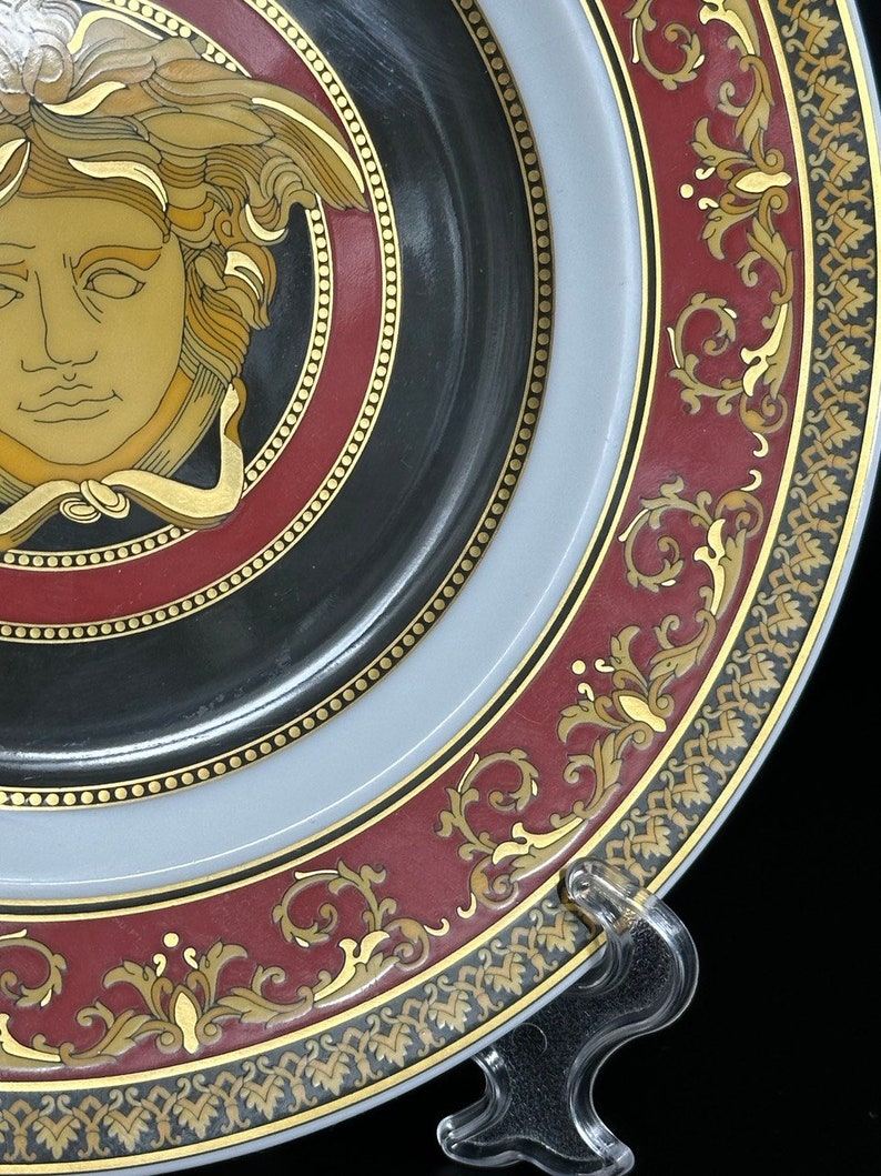Puede incluir: Plato decorativo con un dise&ntilde;o de cabeza de Medusa en oro y marr&oacute;n sobre un fondo negro. El plato presenta un borde burdeos con adornos dorados y un borde gris claro. Un soporte de pl&aacute;stico transparente sostiene el plato.