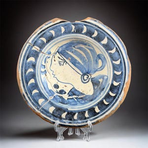 May include: Antique ceramic plate with a blue and white design. The plate features a profile of a person with a head covering. The rim has a repeating pattern of blue and white shapes. The plate is cracked and chipped, showing signs of age.