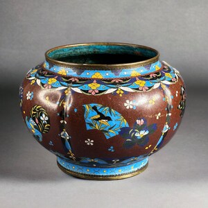 Può includere: Un vaso rotondo in cloisonné marrone con un'ampia apertura e una fascia decorativa di motivi floreali blu, gialli e rosa. Il vaso presenta immagini di uccelli e fiori. Il bordo e la base hanno un bordo blu e oro.