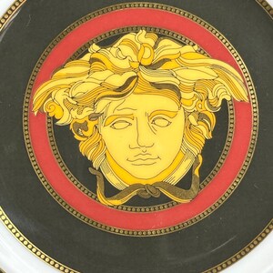 Puede incluir: Un plato decorativo con un dise&ntilde;o central de cabeza de Medusa en oro y amarillo sobre un fondo negro. El plato tiene un borde rojo y un borde blanco con detalles dorados. El dise&ntilde;o del plato es de estilo cl&aacute;sico y elegante.