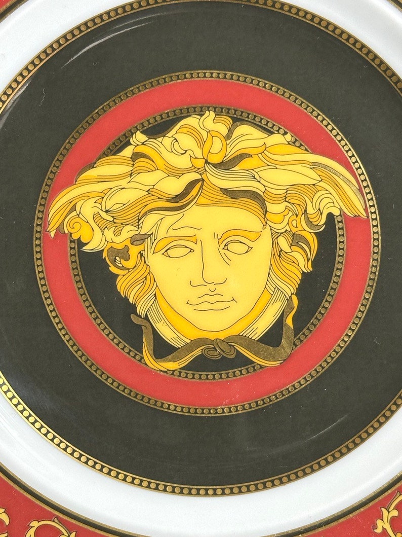 Puede incluir: Un plato decorativo con un dise&ntilde;o central de cabeza de Medusa en oro y amarillo sobre un fondo negro. El plato tiene un borde rojo y un borde blanco con detalles dorados. El dise&ntilde;o del plato es de estilo cl&aacute;sico y elegante.