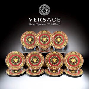 Op de afbeelding: Een set van 12 decoratieve borden, elk met een diameter van 31 cm. De borden hebben een rood en goudkleurig ontwerp met een centraal zwart medaillon en een gouden embleem. De borden zijn op een donkere ondergrond geplaatst.
