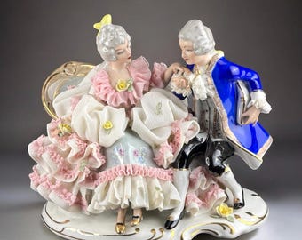May include: A porcelain figurine depicting a couple in 18th-century attire. The woman wears a pink and white dress with ruffles, and the man is in a blue coat. The base is white with gold trim, and the figurine has a romantic theme.