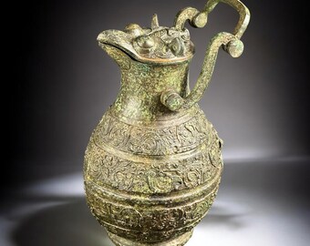 May include: An antique bronze pitcher with a detailed dragon-like spout and handle. The vessel features intricate relief patterns and a lid. The pitcher has a weathered, aged appearance, with a green patina. A decorative piece.