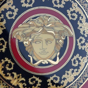Op de afbeelding: Decoratieve plaat met een Medusa-hoofdontwerp. De plaat heeft een zwarte achtergrond met gouden en rode accenten. Het Medusa-hoofd staat centraal, omcirkeld door een rode ring en gouden krullen. De buitenrand heeft een gouden rand.
