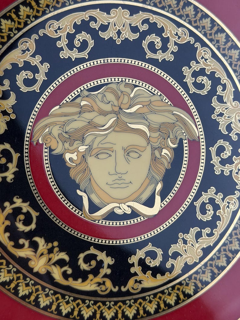 Op de afbeelding: Decoratieve plaat met een Medusa-hoofdontwerp. De plaat heeft een zwarte achtergrond met gouden en rode accenten. Het Medusa-hoofd staat centraal, omcirkeld door een rode ring en gouden krullen. De buitenrand heeft een gouden rand.