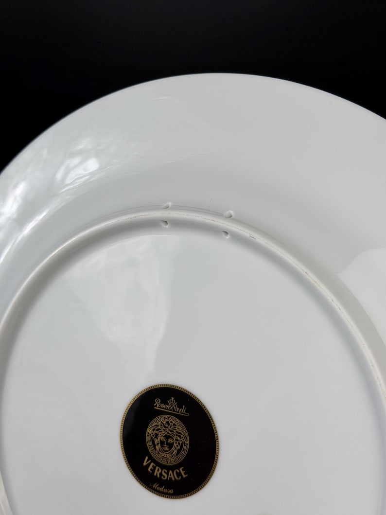 Op de afbeelding: Witte porseleinen bord met een zwart en goud Versace Medusa-logo. Het bord heeft een glanzende afwerking en een licht verhoogde rand. Het Rosenthal-logo is ook zichtbaar.