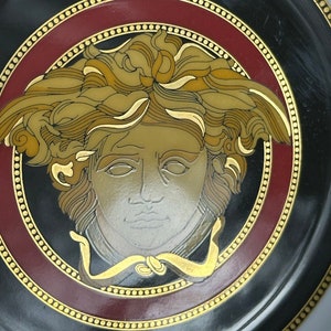Puede incluir: Un plato decorativo negro con un dise&ntilde;o central de cabeza de Medusa en tonos dorados, beige y plateados. El plato tiene un borde burdeos y dorado, con un ribete dorado punteado. El plato es un art&iacute;culo decorativo.