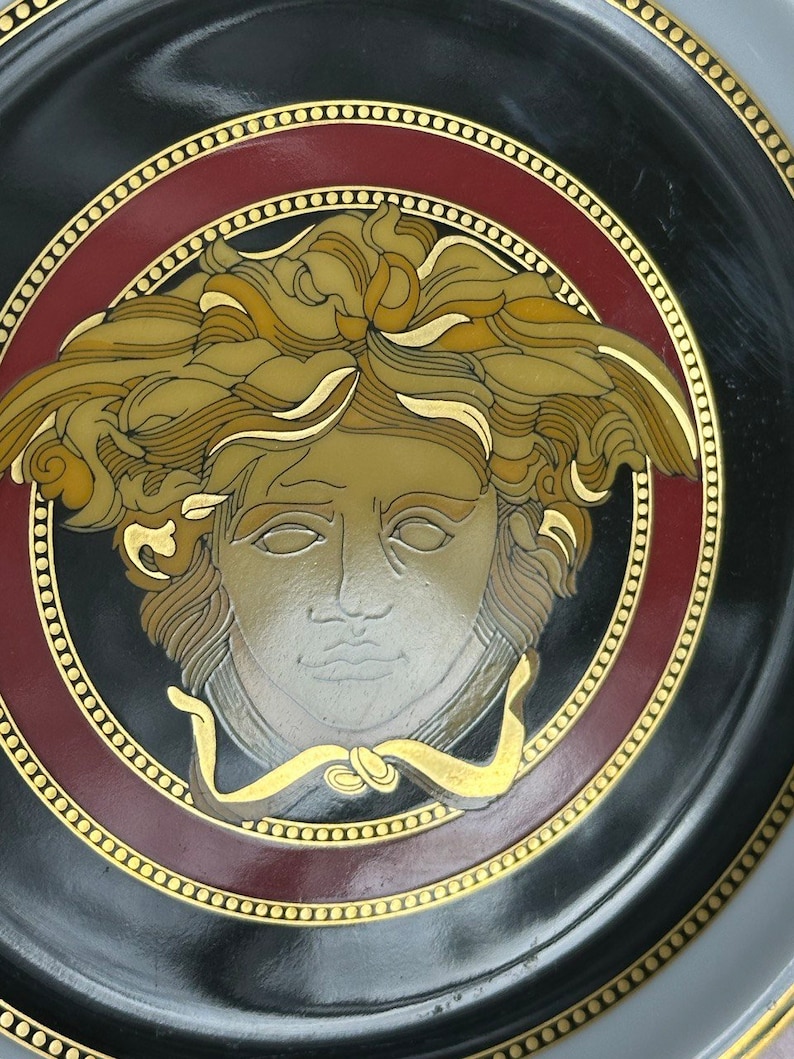 Puede incluir: Un plato decorativo negro con un dise&ntilde;o central de cabeza de Medusa en tonos dorados, beige y plateados. El plato tiene un borde burdeos y dorado, con un ribete dorado punteado. El plato es un art&iacute;culo decorativo.